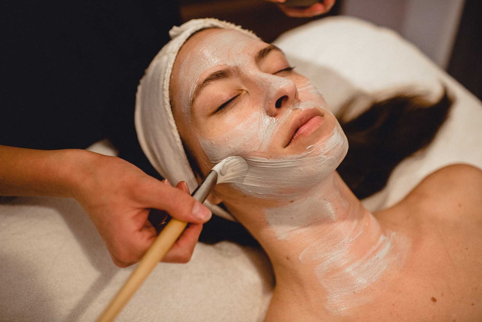 Woman receiving a facial treatment with a brush applying a white mask at a spa.