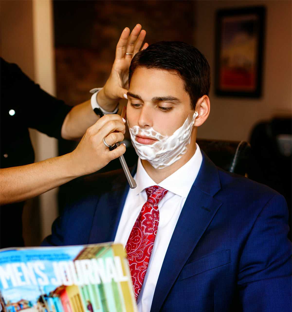 Man in suit getting shave while reading a magazine.