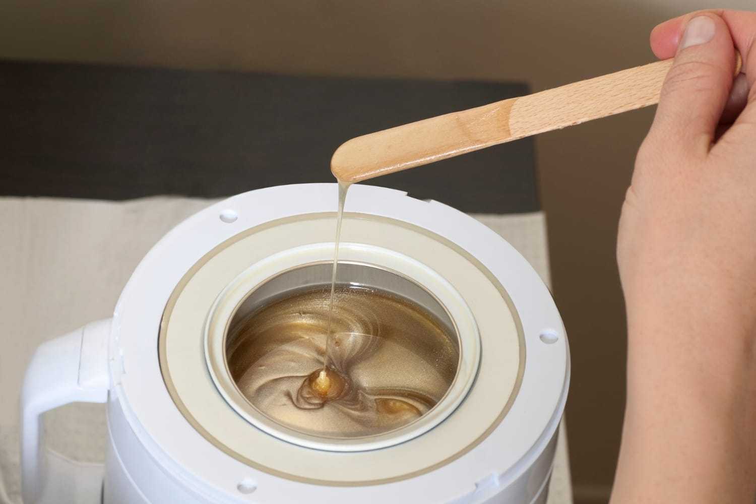 Mixing honey-colored wax in a white heater with a wooden spatula.