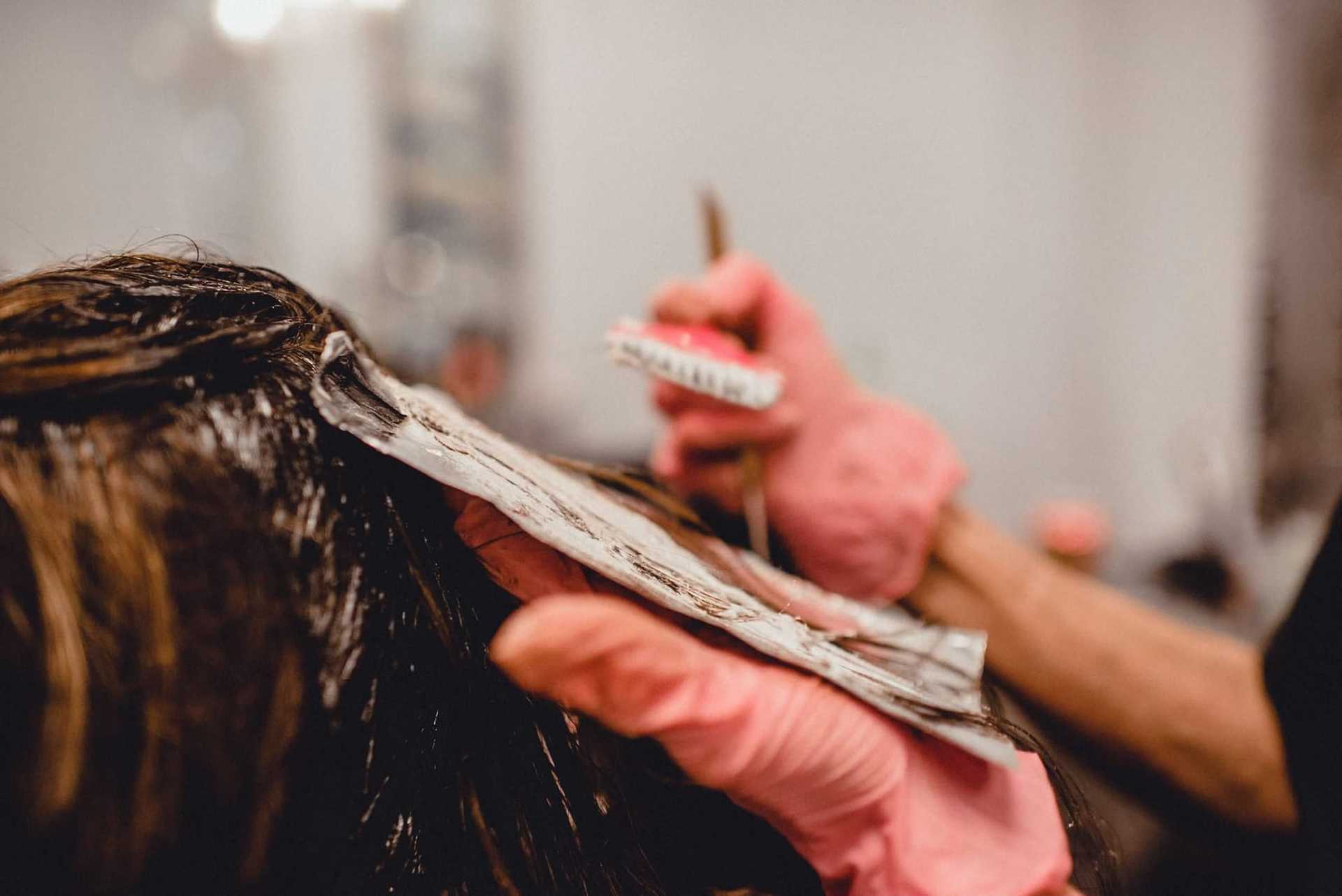 Hairdresser applying color to client's hair using a brush in a salon, wearing pink gloves.